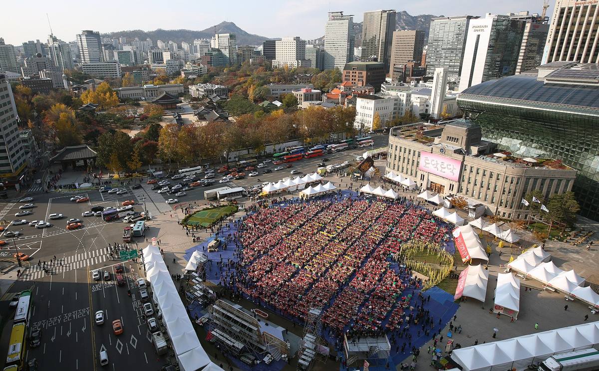 An aerial view of Seoul Plaza in November 2014 filled with rows of people in red aprons making kimchi at long tables, Seoul City Hall and Deoksugung's autumn trees behind them.