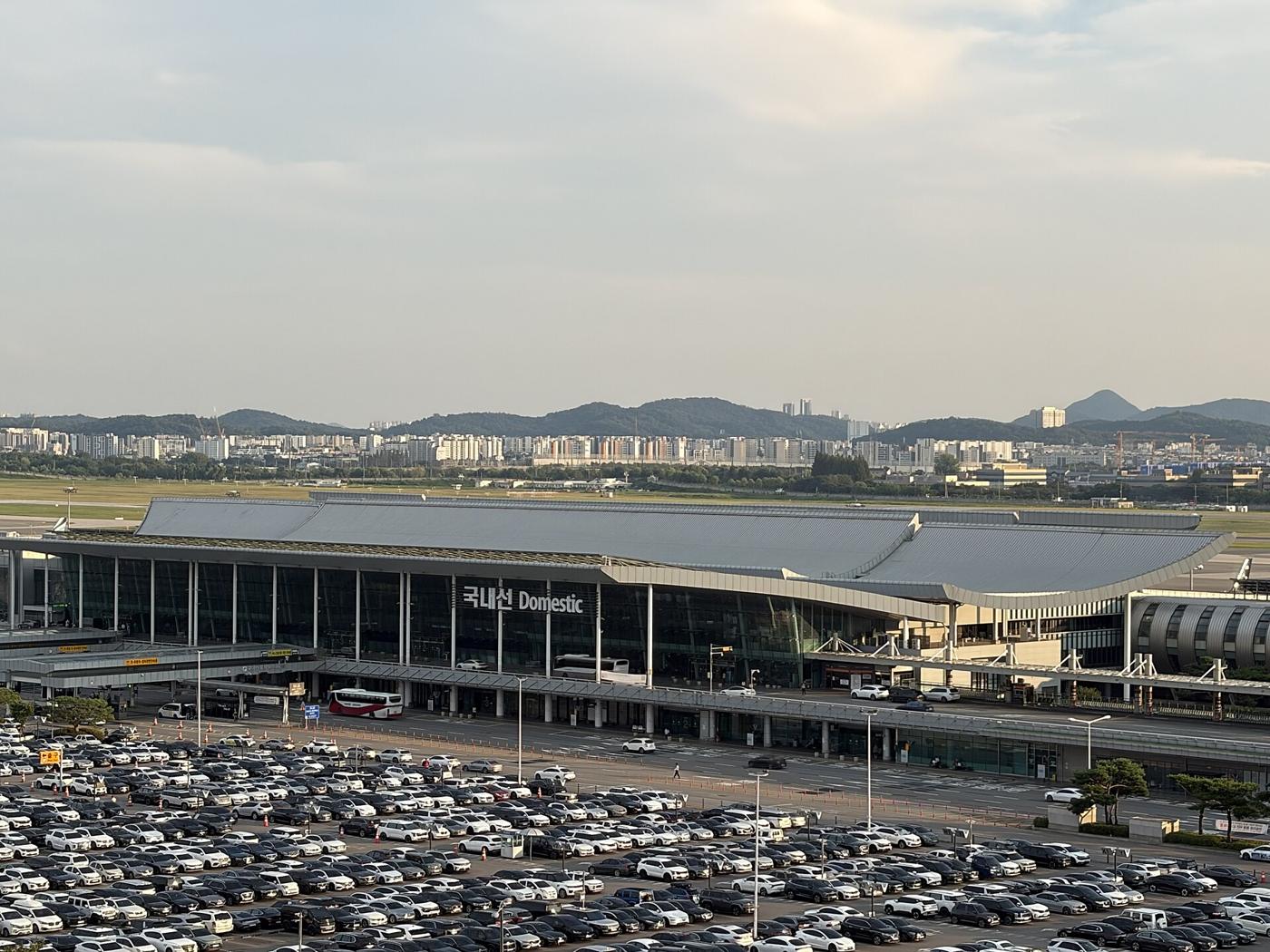 The domestic terminal of Gimpo International Airport seen from a distance, its long curved roof and a large "국내선 Domestic" sign above the glass front, with a full car park in the foreground.