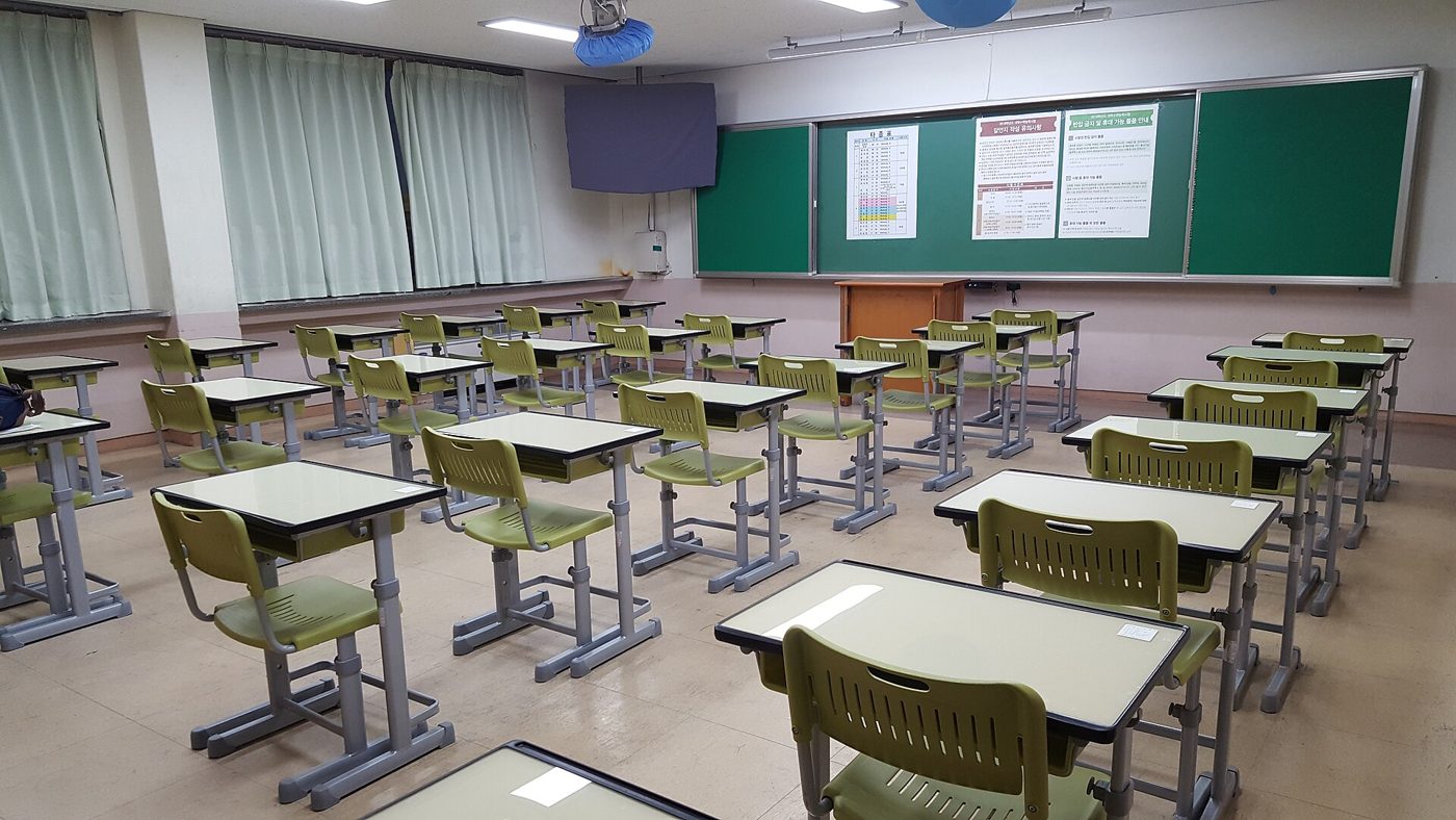 An empty Korean classroom prepared as an exam room: single desks spaced in rows with green chairs, and notices and a timetable pinned to the green board at the front.