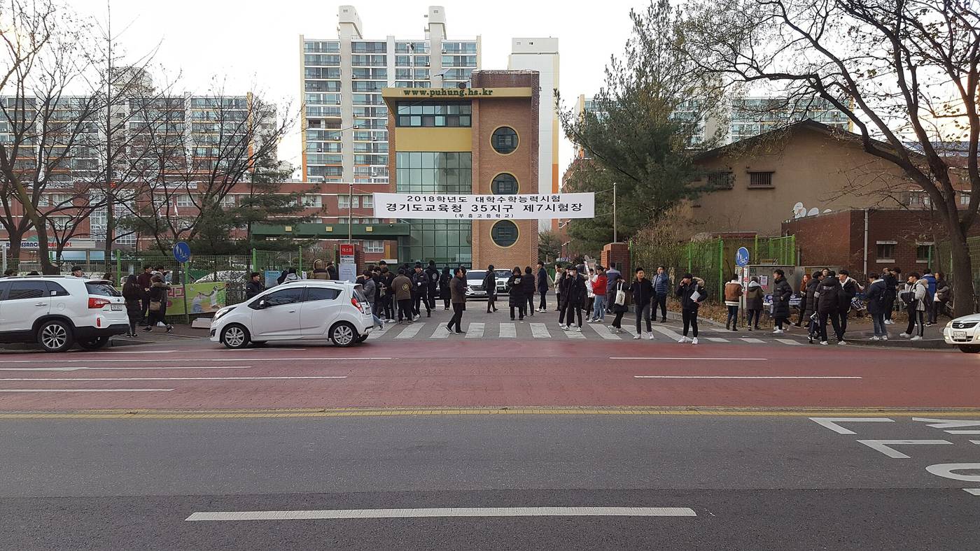 Students in dark winter jackets with backpacks walking out of a school gate on exam day, crossing the road while a few parents wait by the kerb, with the test-site banner and apartment blocks behind them.