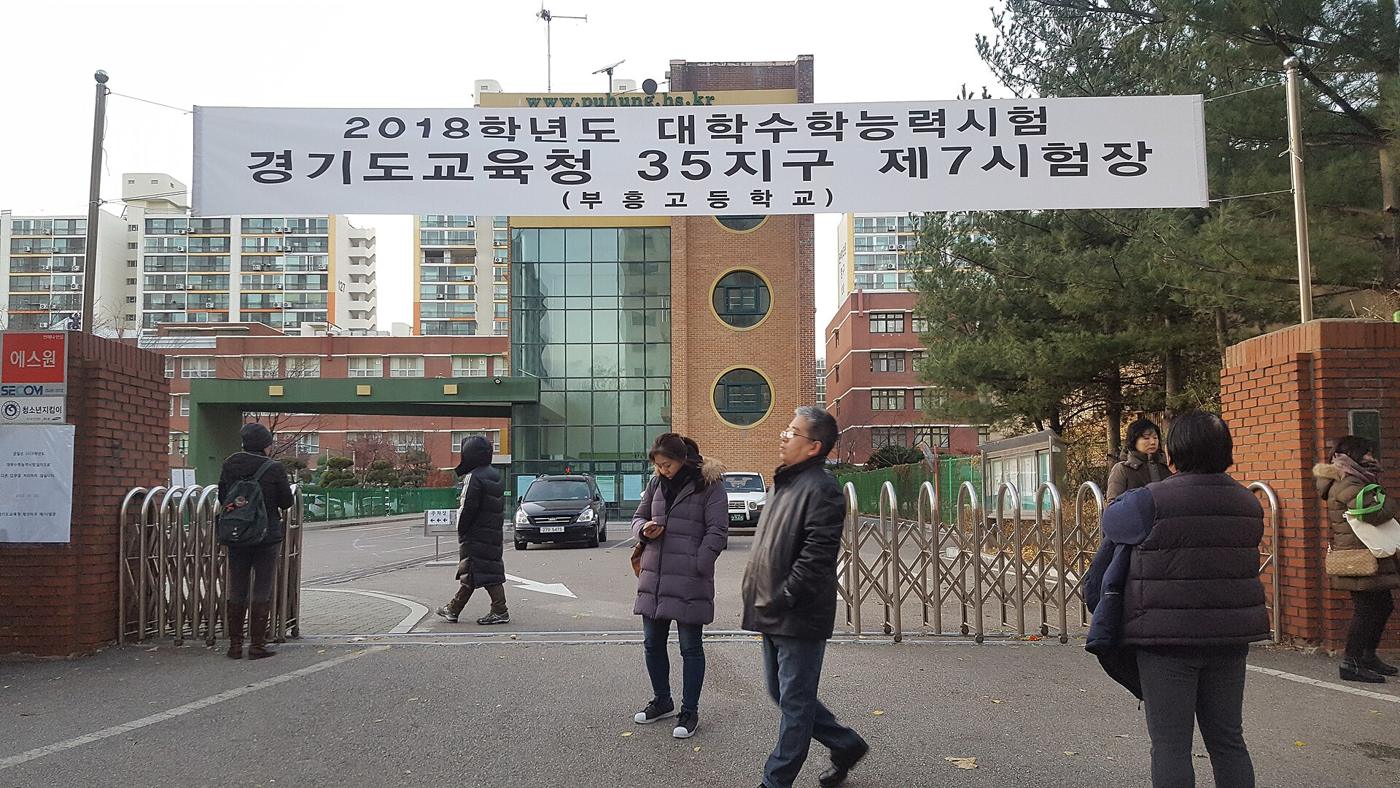A white banner reading "2018학년도 대학수학능력시험 경기도교육청 35지구 제7시험장 (부흥고등학교)" hangs over the open gate of Puhung High School, with a few parents in winter coats waiting outside and apartment blocks behind the school.