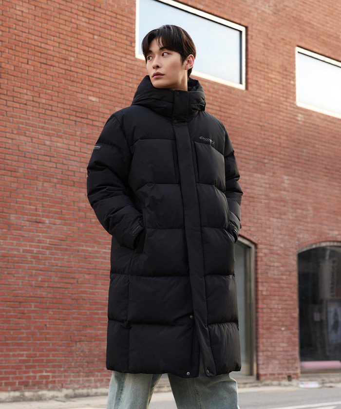 Discovery Expedition's Lester G goose-down long padding in black, a hooded, knee-length quilted parka worn over jeans in front of a brick wall.