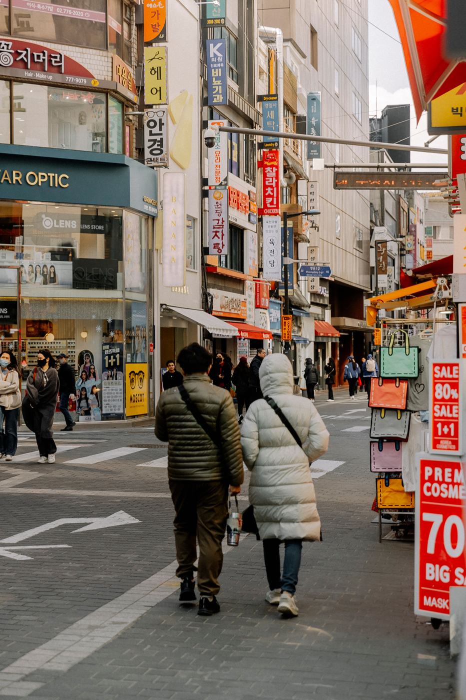 A couple walking down a Seoul shopping street in winter; the woman wears a long cream padded coat that reaches to her knees, with shop signs and cosmetics sale boards along the street.