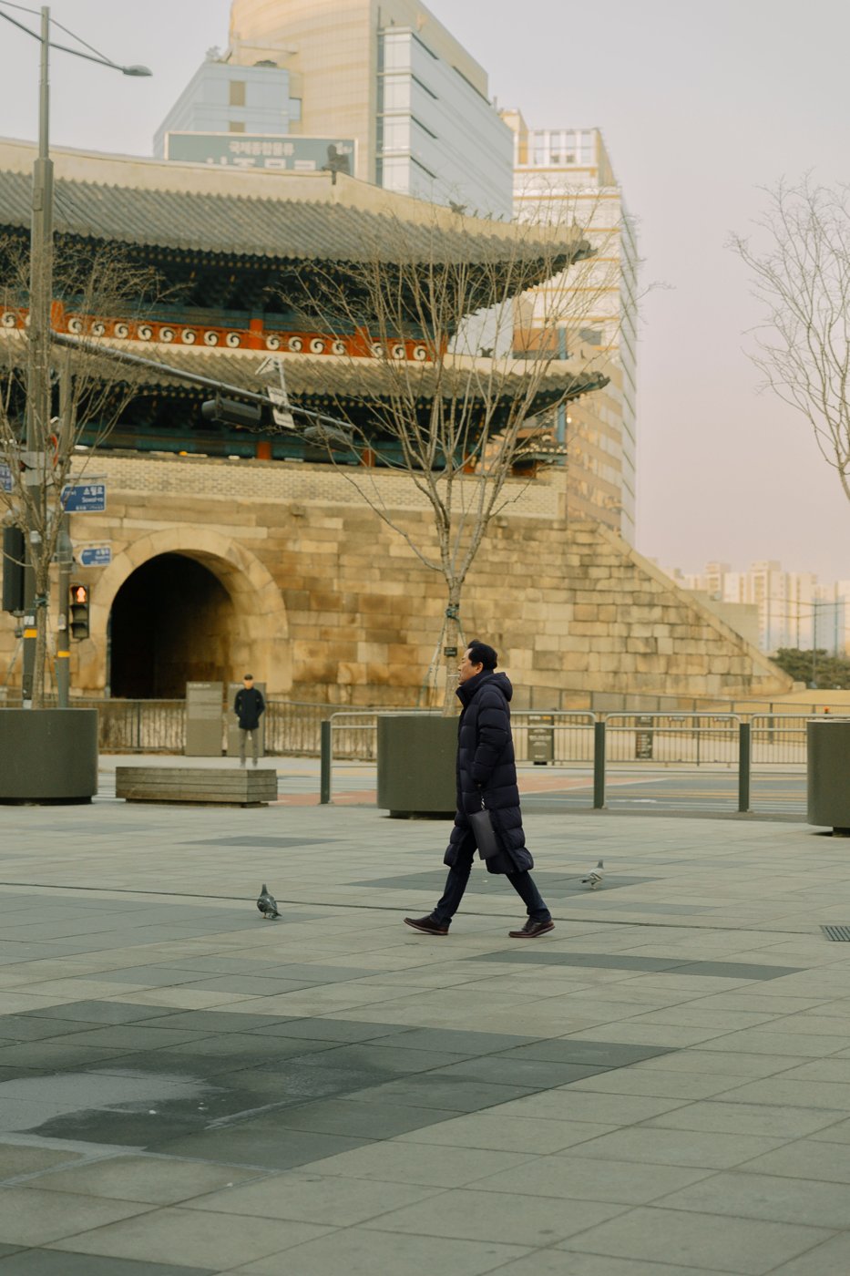A man in a long black padded coat walking across an empty plaza in front of Sungnyemun (Namdaemun), Seoul's South Gate, on a cold, clear late-winter afternoon.