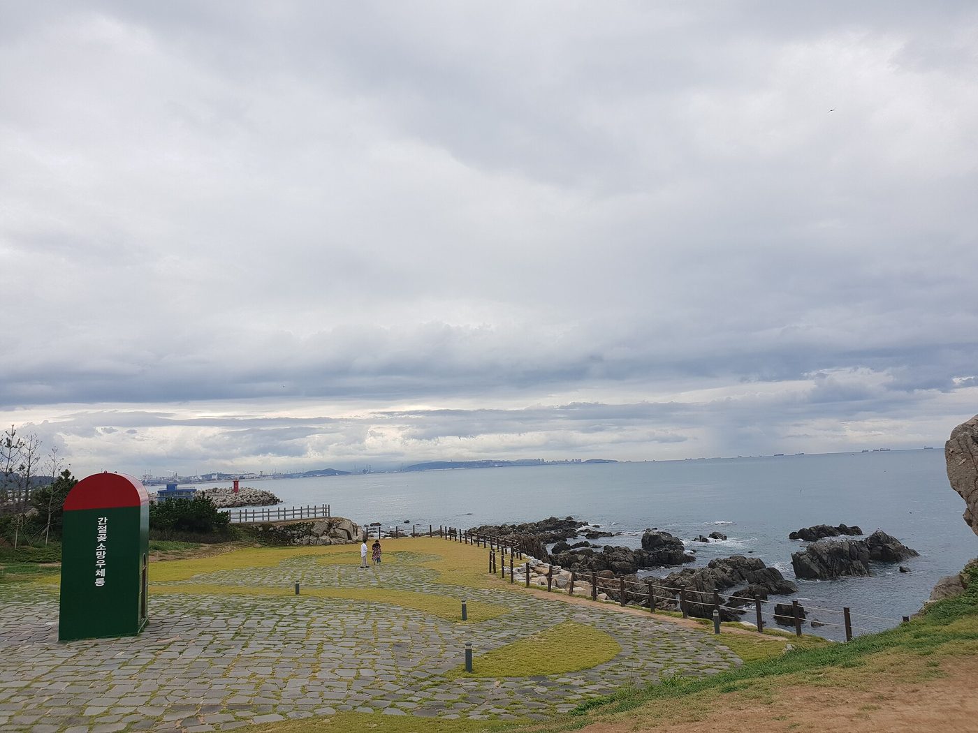The grassy headland at Ganjeolgot under a grey sky, with a tall red-and-green post box labelled 간절곶 소망우체통 on a stone-paved lawn and a wooden fence running down to dark rocks and the sea.