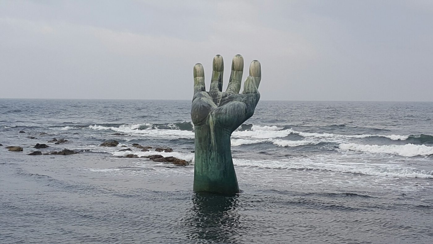 A large bronze hand with outstretched fingers rising out of grey sea water, waves breaking around its wrist, under an overcast sky at Homigot.
