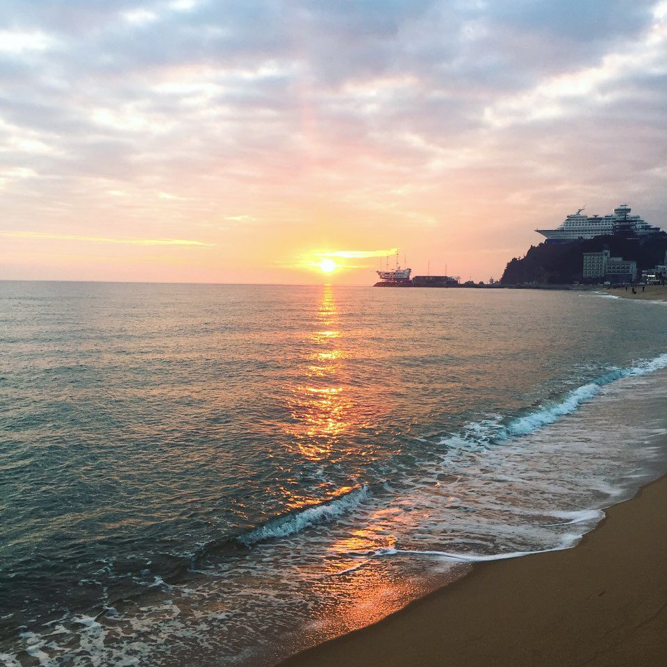 The sun low over a calm sea at Jeongdongjin, with a bright path of light on the water, waves breaking on the sand in front, and the ship-shaped Sun Cruise resort on the headland at right.