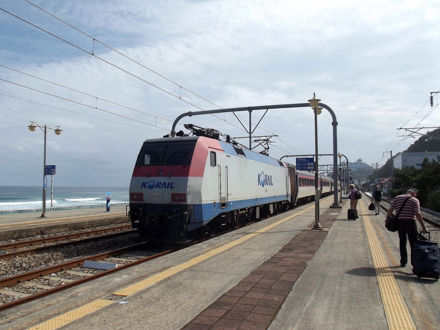 A Korail electric locomotive pulling a passenger train along the platform of Jeongdongjin Station, with the sea and breaking waves just beyond the far track and a cruise-ship-shaped hotel on the cliff in the distance.