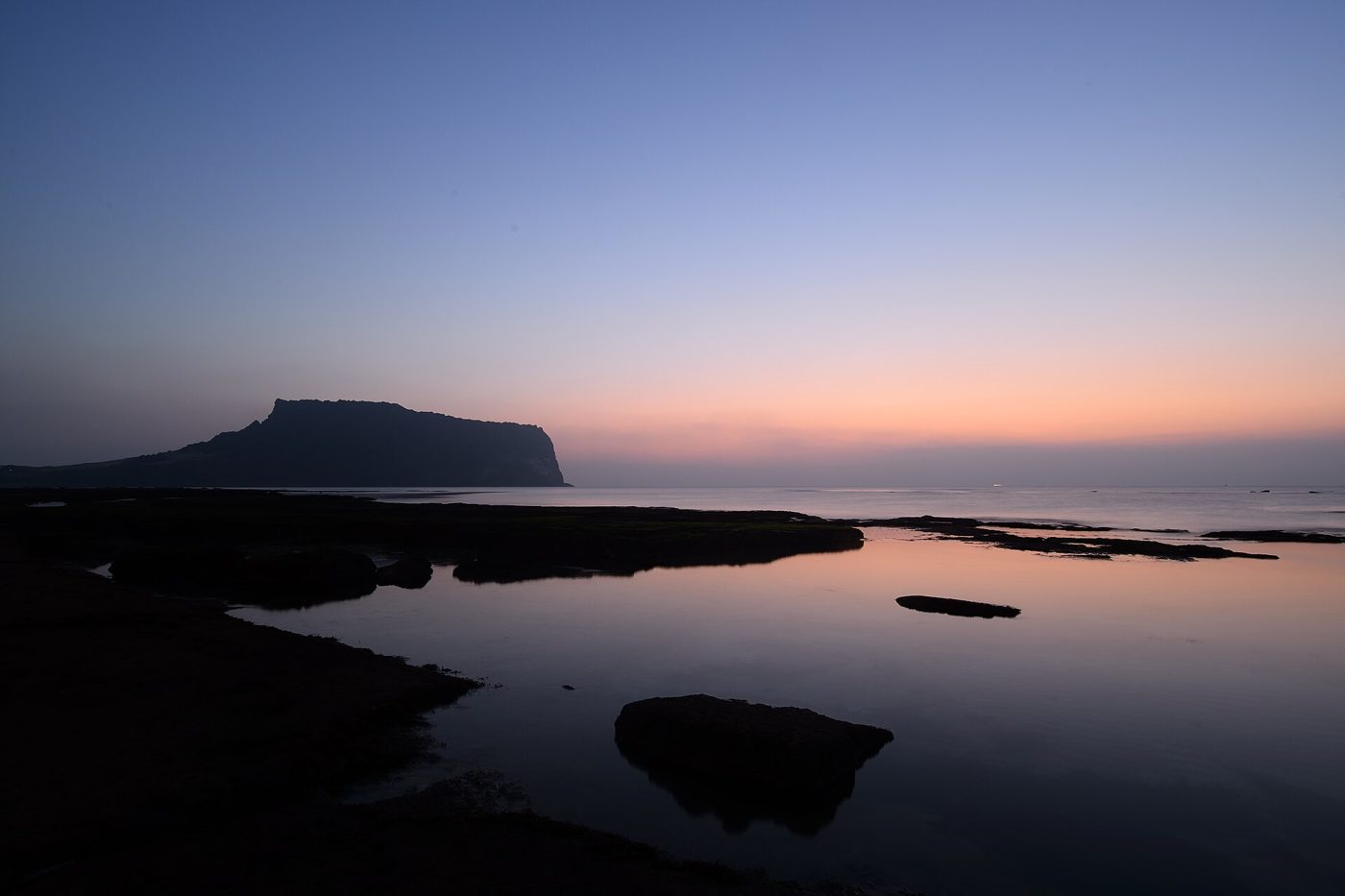 The dark silhouette of Seongsan Ilchulbong crater against a pale pink and blue dawn sky, with the pink sky reflected in a calm tidal pool on the black rocks in front.