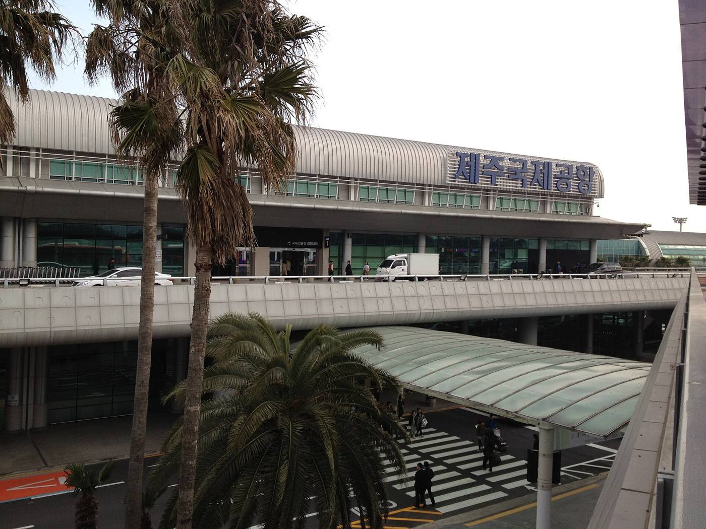 The front of Jeju International Airport with the blue sign 제주국제공항 on the roof, palm trees in the foreground and travellers crossing the road to the covered bus and taxi area below the departures ramp.