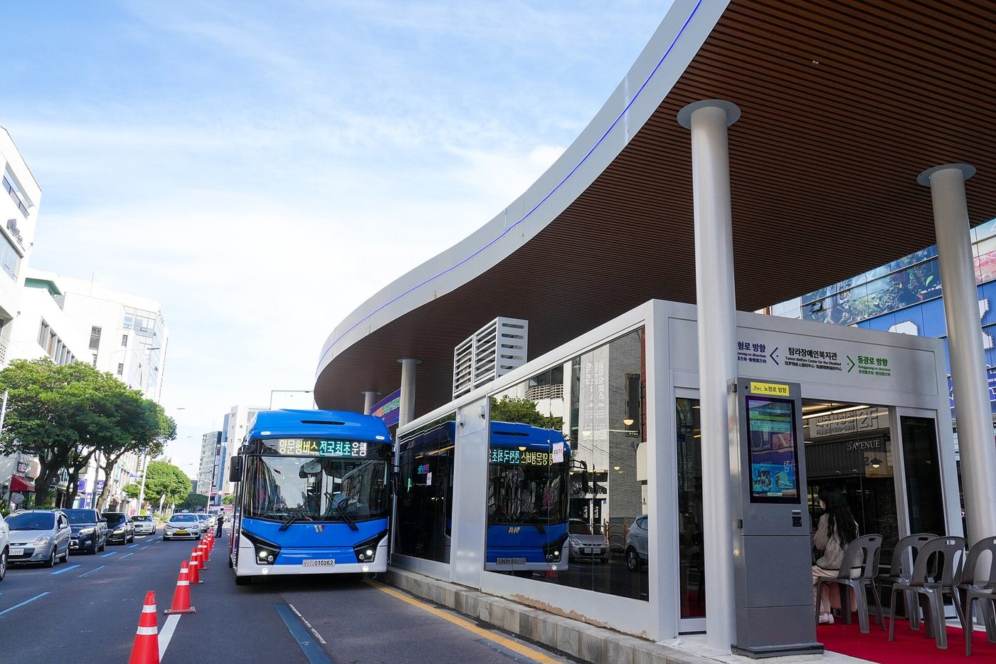 Two blue Jeju city buses pulled up at a new island bus stop with a curved wooden canopy, glass waiting room and a digital information screen, on a sunny street in Jeju City; the front bus's sign reads 양문형버스 전국최초운행.