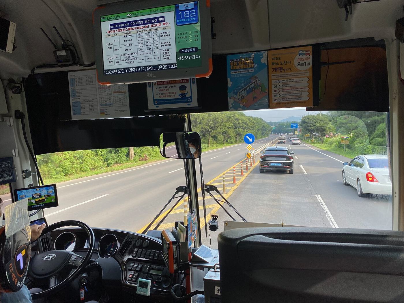 The view from behind the driver of Jeju express bus 182: a route screen showing "182" and the next stop, a notice reading "제주도 버스 노선이 2024년 8월 1일 개편됩니다", and a four-lane road lined with trees ahead.