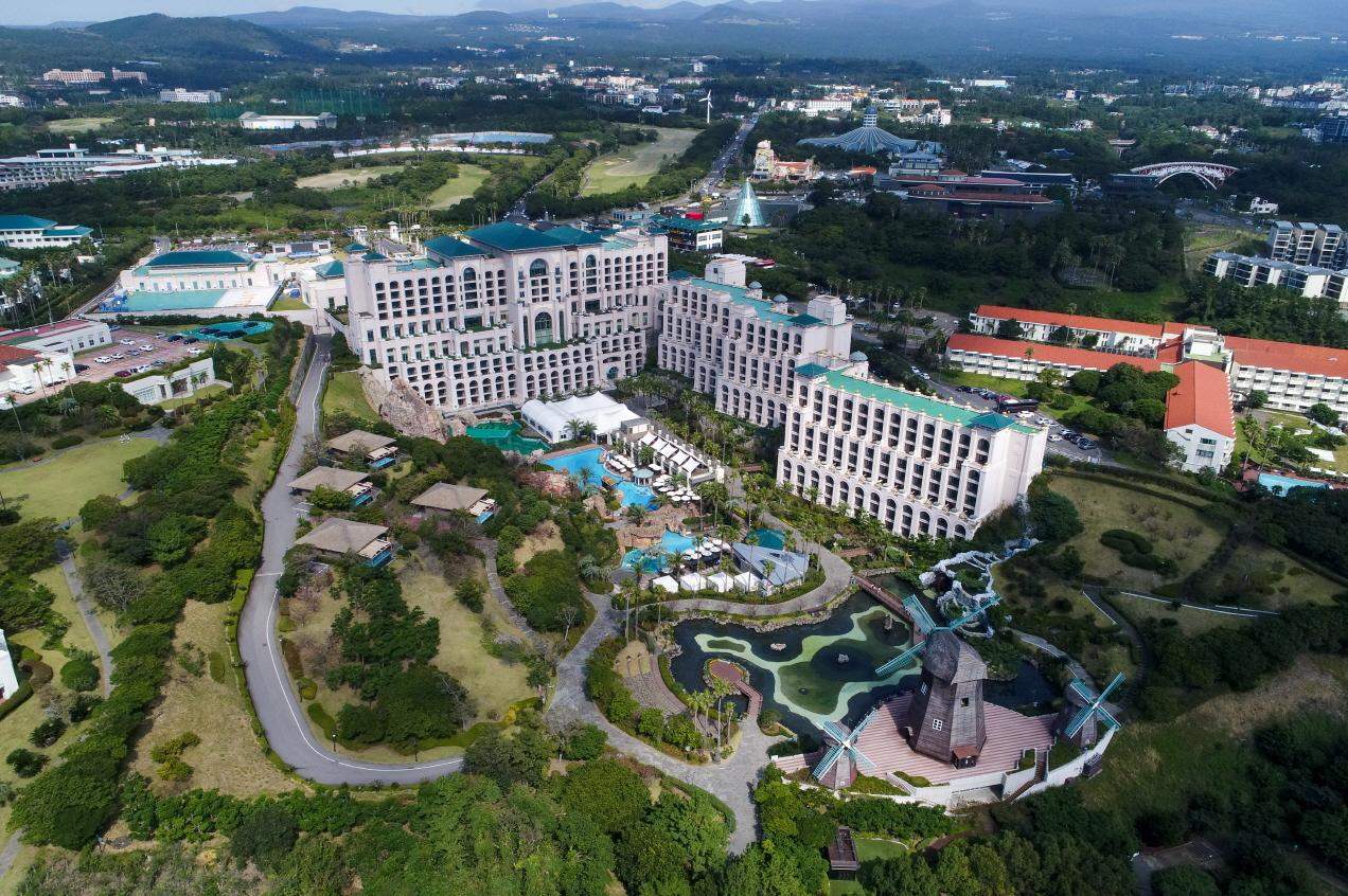 An aerial view of Lotte Hotel Jeju in the Jungmun resort: a large cream-coloured hotel with green roofs, pools and gardens, a pond with Dutch-style windmills in the foreground, and the island's hills behind.