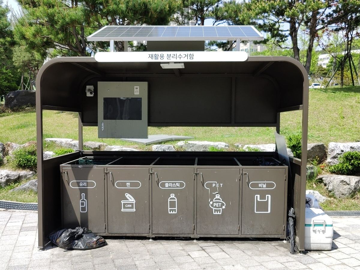 A solar-roofed recycling station in Korea with five labelled bays: 유리 glass, 캔 cans, 플라스틱 plastic, a separate bay for transparent PET bottles, and 비닐 vinyl.