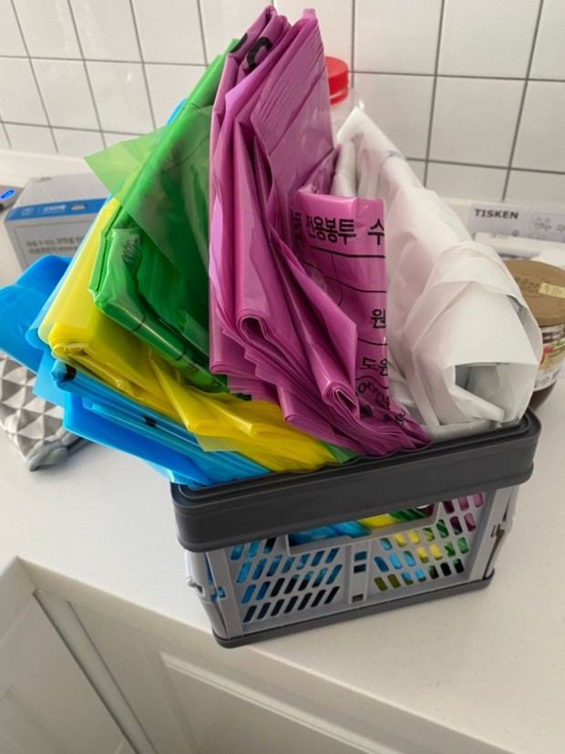 A plastic basket on a kitchen counter in Korea holding folded waste bags in blue, yellow, green, pink and white, with Korean printing on them.