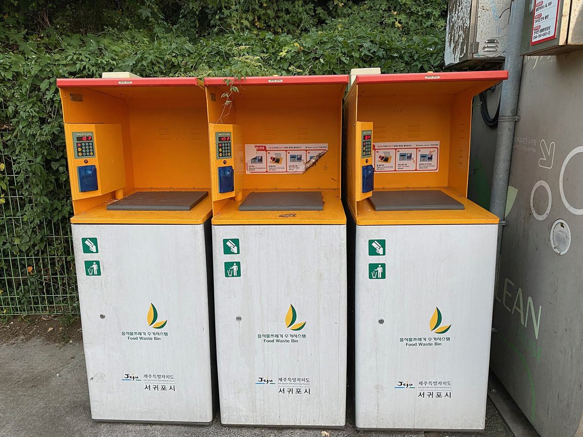 Three white and orange RFID food-waste bins at a roadside in Seogwipo, Jeju, each with a keypad, a card reader and a weight display, labelled 음식물쓰레기 수거시스템.