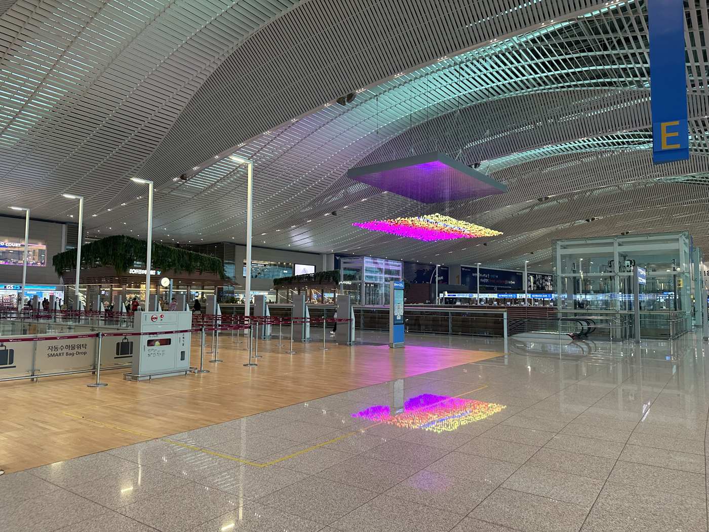 The Terminal 2 departures hall at Incheon International Airport, with self bag-drop machines on a wooden floor, check-in row E, and a light sculpture hanging from the ceiling.