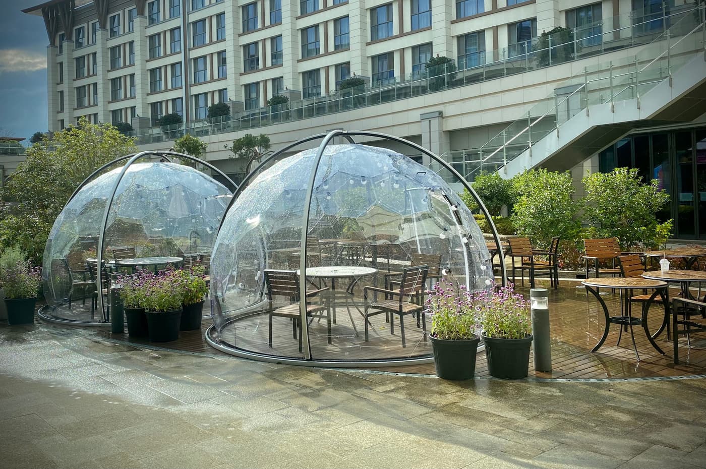 A terrace with two transparent bubble domes holding tables and chairs, in front of a multi-storey resort hotel building.