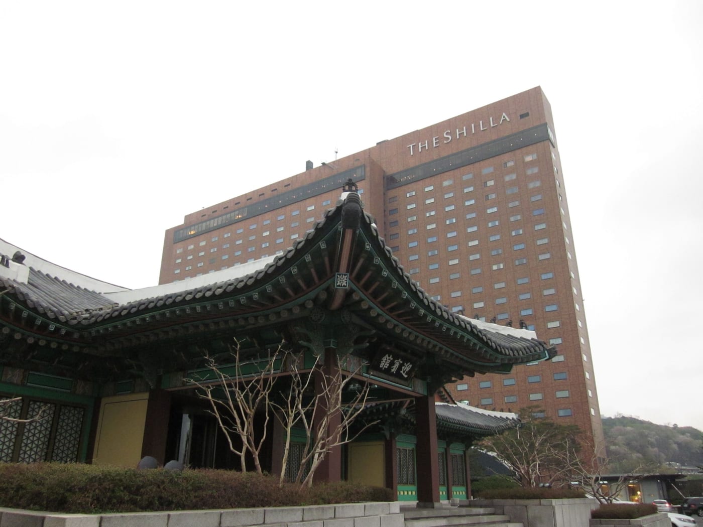 The Shilla Seoul hotel tower behind the curved tiled roof of a traditional Korean building at its entrance.