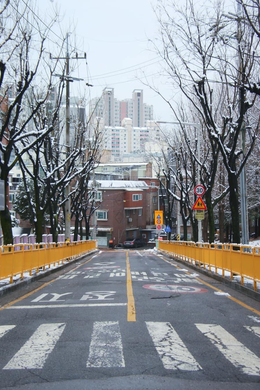A snowy residential street in Korea in a school zone, with a 30 km/h sign and Korean road markings, a red-brick villa building at the end of the road and taller apartment towers behind it.