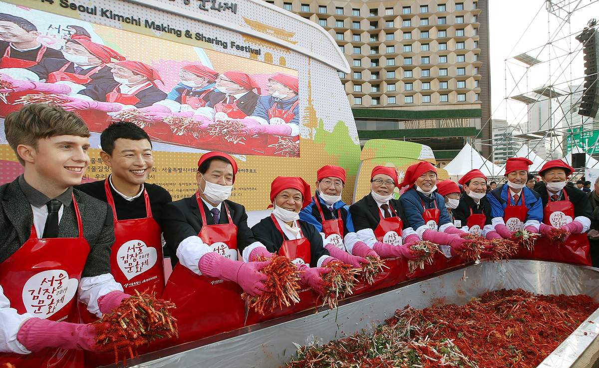 A row of guests in red aprons and pink rubber gloves, most in red headscarves, hold up handfuls of red radish-and-chilli kimchi filling over a long steel trough at the 2014 Seoul Kimjang Culture Festival in Seoul Plaza.