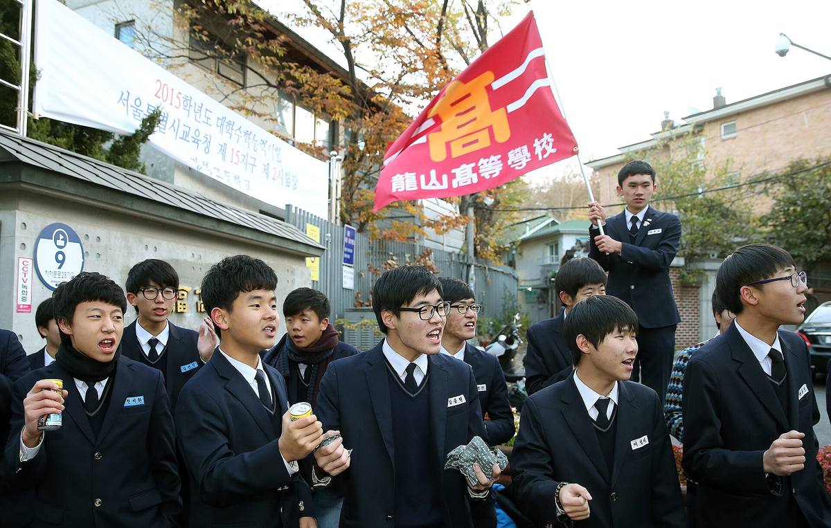 Students in navy school uniforms cheer outside a Seoul exam site on Suneung morning in November 2014, under a banner for the 2015 exam and a red flag reading 龍山高等學校, Yongsan High School.