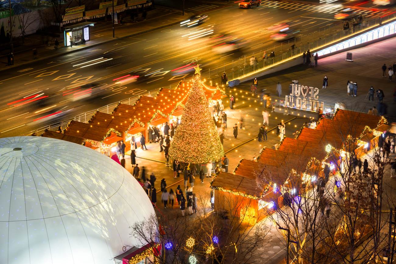 The Gwanghwamun Market in Seoul at night seen from above: a lit Christmas tree with a star surrounded by red-roofed wooden market huts strung with lights, a white dome and traffic on Sejong-daero beside it.