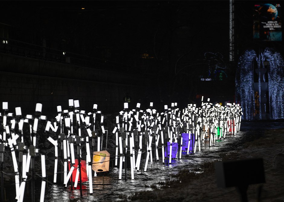 White light-stick sculptures standing in the shallow water of Cheonggyecheon stream at night during the 2025 Seoul Lantern Festival, with an arch of white lights behind.