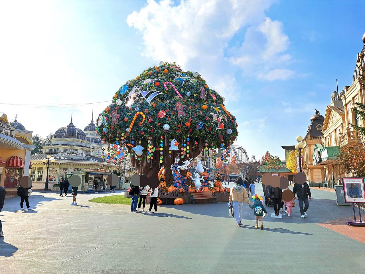 Everland's Magic Tree on the main avenue near the entrance, decorated for Halloween with pumpkins, ghost figures, candy canes and spider webs, with a crowd of visitors under a blue sky and Western-style park buildings on both sides.