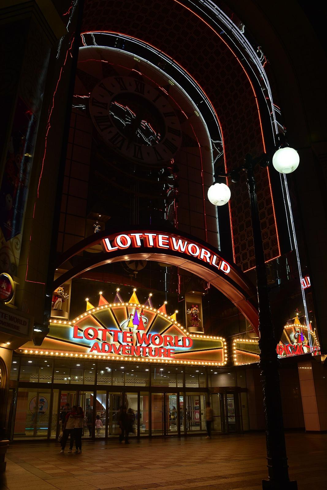 The Lotte World Adventure entrance at night, with the illuminated LOTTE WORLD arch, a large clock face above it, and a marquee sign reading LOTTE WORLD ADVENTURE over the glass entrance doors.