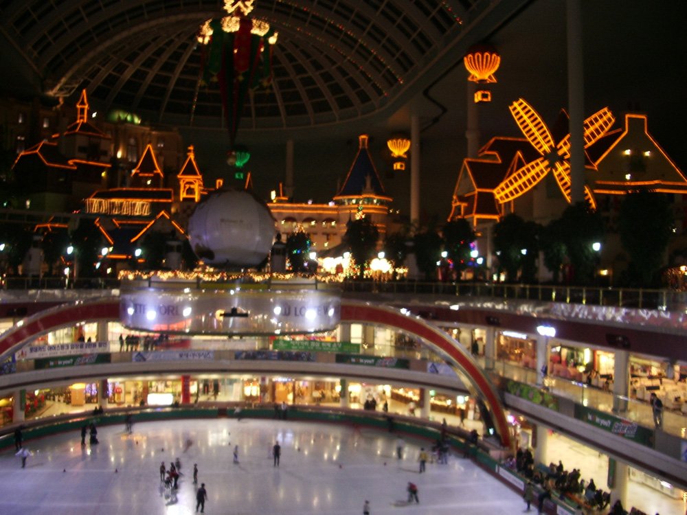 The interior of Lotte World Adventure at night, with the ice rink on the lowest level, ringed shopping and dining floors around it, and illuminated buildings and a windmill under the glass dome above.
