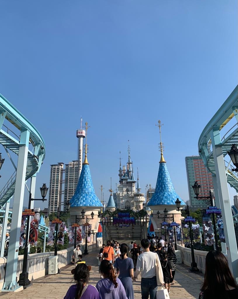 The bridge to Lotte World's outdoor Magic Island, lined with Halloween banners, leading to the white Magic Castle with blue cone-shaped towers, with the Gyro Drop tower and apartment blocks behind.