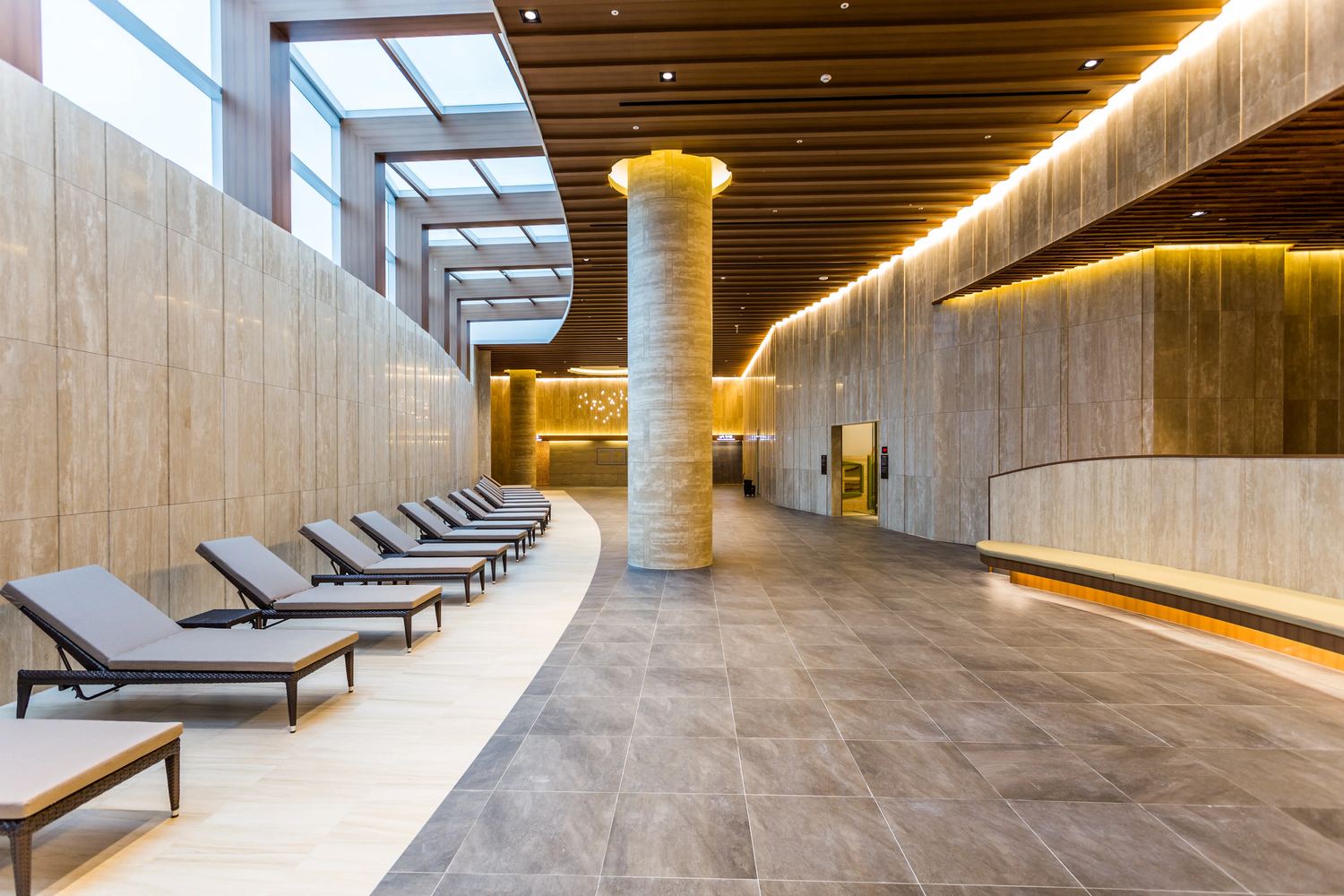 Aquafield Hanam's jjimjil spa hall: a curved row of grey sun loungers along a stone wall under skylights, a wide grey-tiled floor, a stone column and a wooden slatted ceiling with warm lighting.