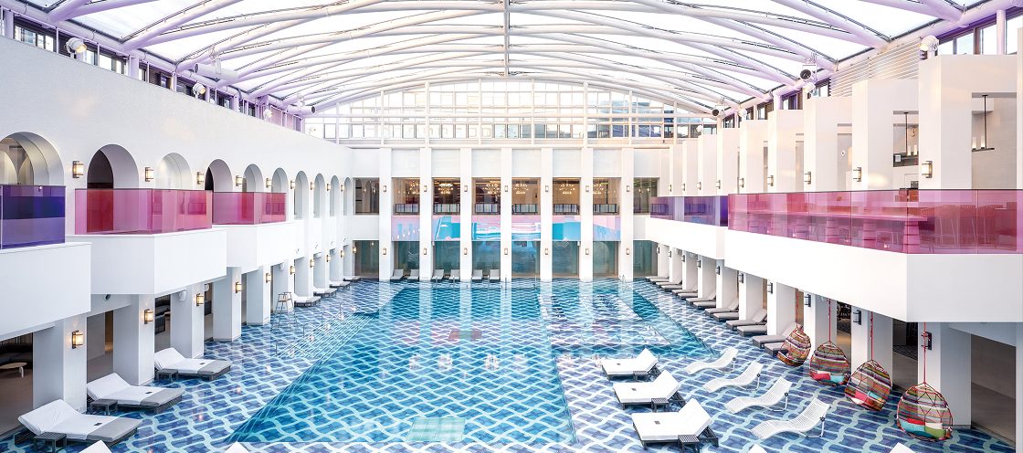 Cimer's indoor pool hall at Paradise City: a large pool with blue and white wave-pattern tiles under a glass barrel roof, white arcades with pink glass balconies, white sun loungers and hanging basket chairs.