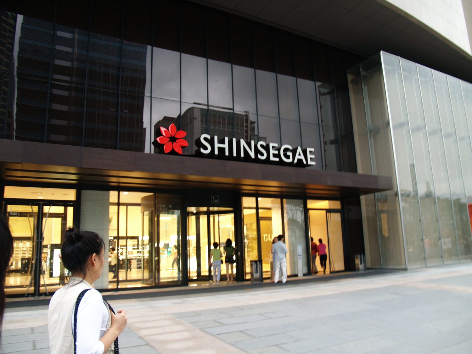 The ground-floor entrance of Shinsegae Centum City in Busan, with the red flower logo and "SHINSEGAE" in white letters above glass doors, and shoppers walking in.