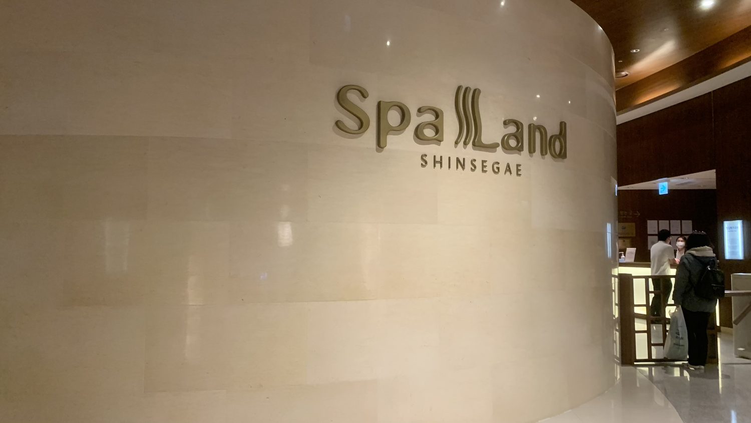The curved stone entrance wall of Spa Land with its gold "Spa Land SHINSEGAE" logo, and the ticket counter on the right with two customers standing at it.