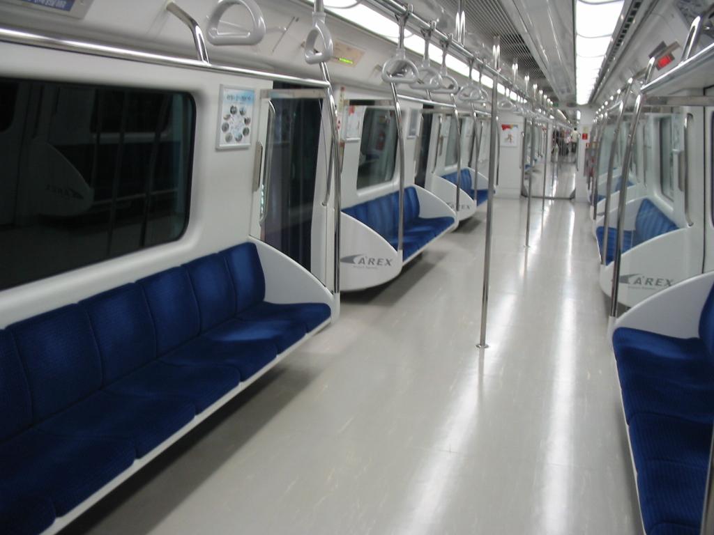 Inside an AREX all-stop commuter train: long blue bench seats along both walls, grab poles and a clear floor for luggage.