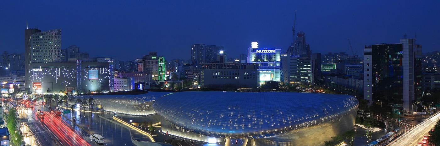 Dongdaemun Design Plaza at night: the long, curved, lit aluminium shell of the DDP in the foreground, with the lights of Dongdaemun's shopping towers and traffic behind.