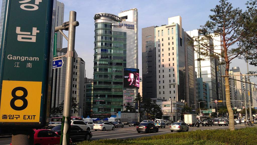 Gangnam Station exit 8: the green station sign with a large number 8, glass office towers and traffic on Gangnam-daero behind it.