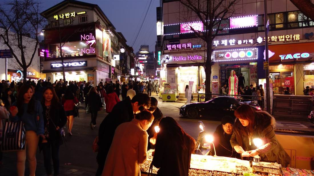 Hongdae at night: crowds walking past cosmetics shops and neon signs while two stallholders sell jewellery under lamps in the foreground.