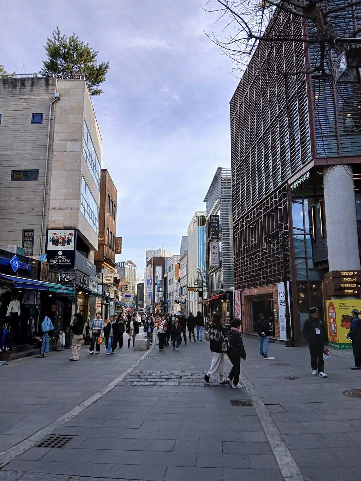 Insadong-gil: a wide pedestrian street paved in grey stone, shops with hanging signs on both sides, people walking toward the camera under a late-afternoon sky.