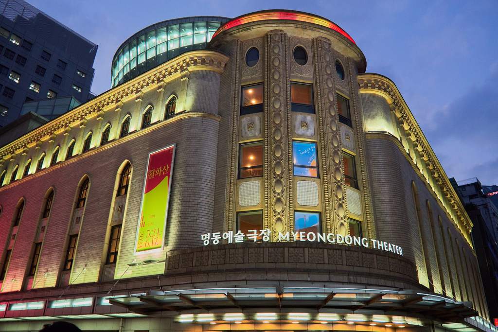 Myeongdong Theater (명동예술극장) at dusk, its curved, lit Art Deco facade with the Korean and English name above the entrance, a theatre banner on the left wall.