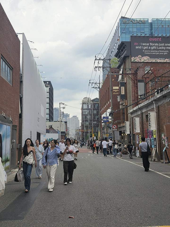 A street in Seongsu-dong: red-brick former factory buildings with shop banners on both sides, shoppers walking down the middle of the road, high-rise apartments in the distance.
