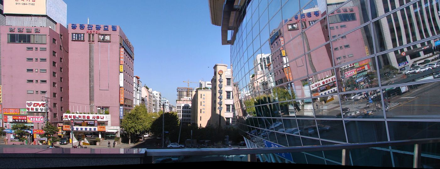 The view north from the main entrance of Busan Station: a wide road lined with older pink and beige hotel and office buildings with Korean signs, and the station's glass wall reflecting the street on the right.