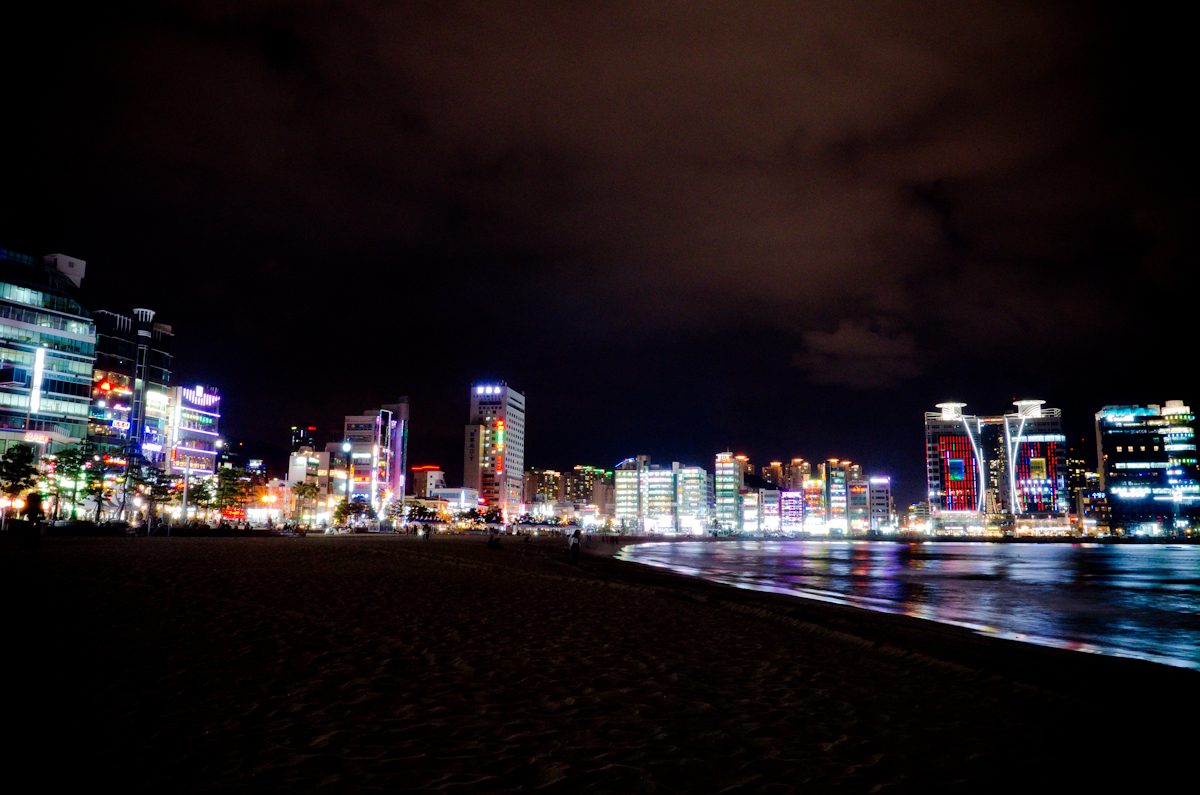 Gwangalli Beach at night: dark sand in the foreground, the curve of the water, and a long row of lit beachfront buildings along the shore.
