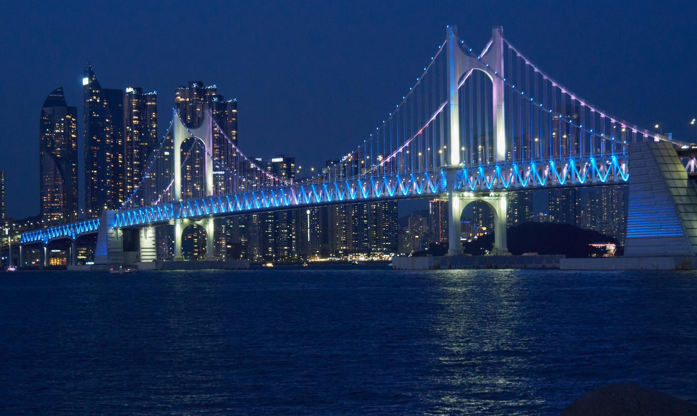 Gwangan Bridge lit blue and white at night over dark water, with the tall residential towers of Haeundae's Marine City lit up behind it.