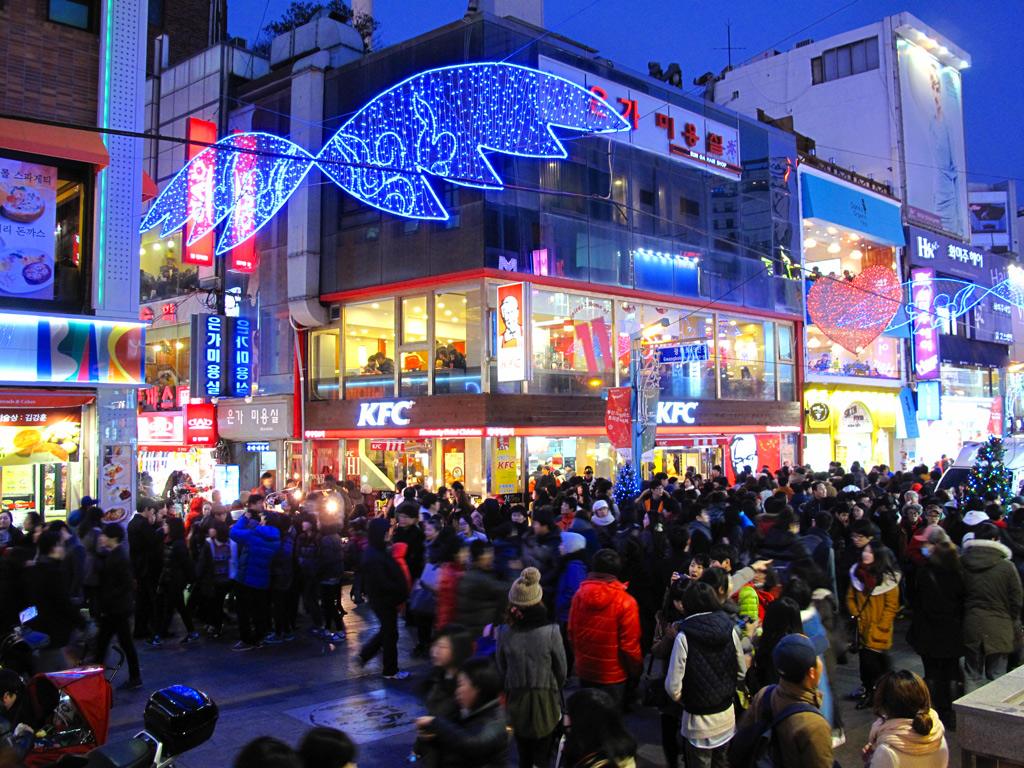 A crowded pedestrian street in Nampo-dong, Busan, in winter, with blue fish-shaped and heart-shaped light sculptures strung over the street and people in padded coats.