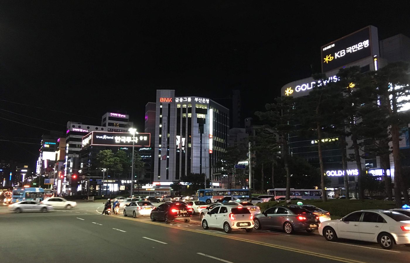 Seomyeon Rotary in Busan at night: a wide road with traffic and city buses, and illuminated office towers with KB Kookmin Bank and BNK Busan Bank signs.