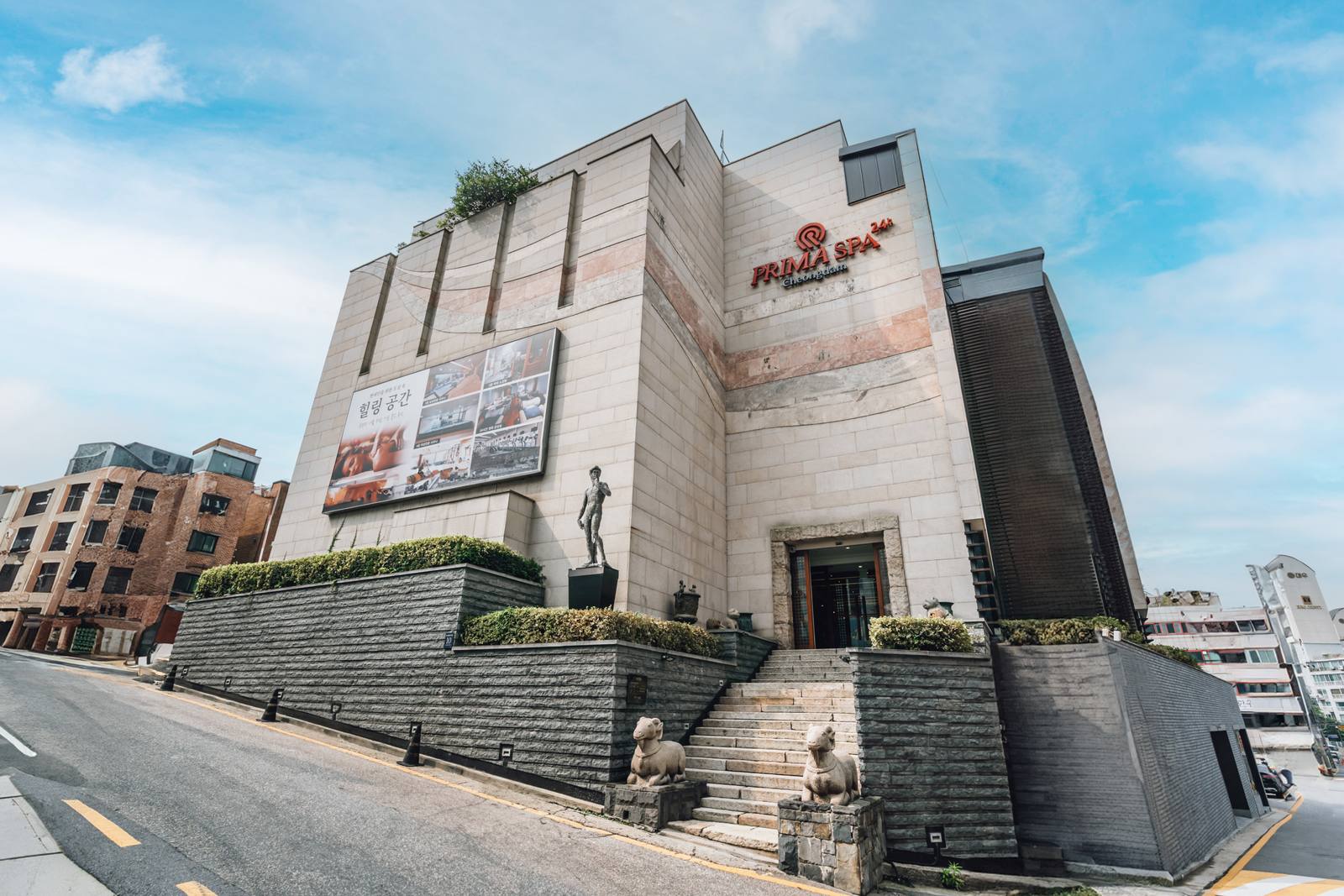 The stone-clad Prima Spa building in Cheongdam with a red PRIMA SPA 24h Cheongdam sign, a wide stone staircase flanked by two stone sheep statues, and a healing-space banner.