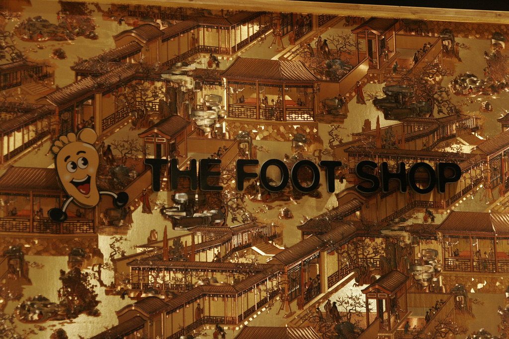 A gold mural of a traditional Chinese town with tiled roofs and wooden balconies, with a cartoon foot mascot and the words THE FOOT SHOP in black letters.